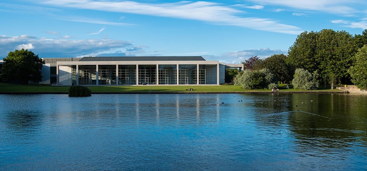 Picture of O'Reilly Hall. It's a long building with pillars pictured from across the UCD lake.