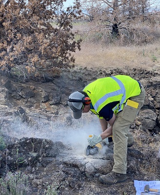 A scientific researcher wearing a high-jacket waistcoat and noise protectors uses an angle grinder to extract lava samples