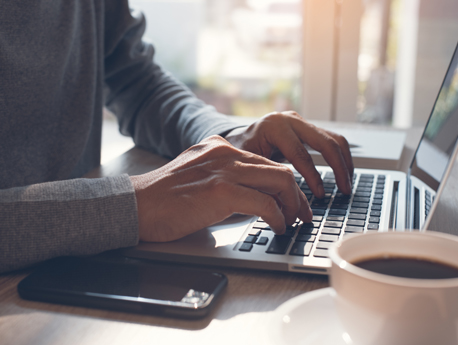 Hands typing at a laptop keyboard, beside a phone and cup of coffee