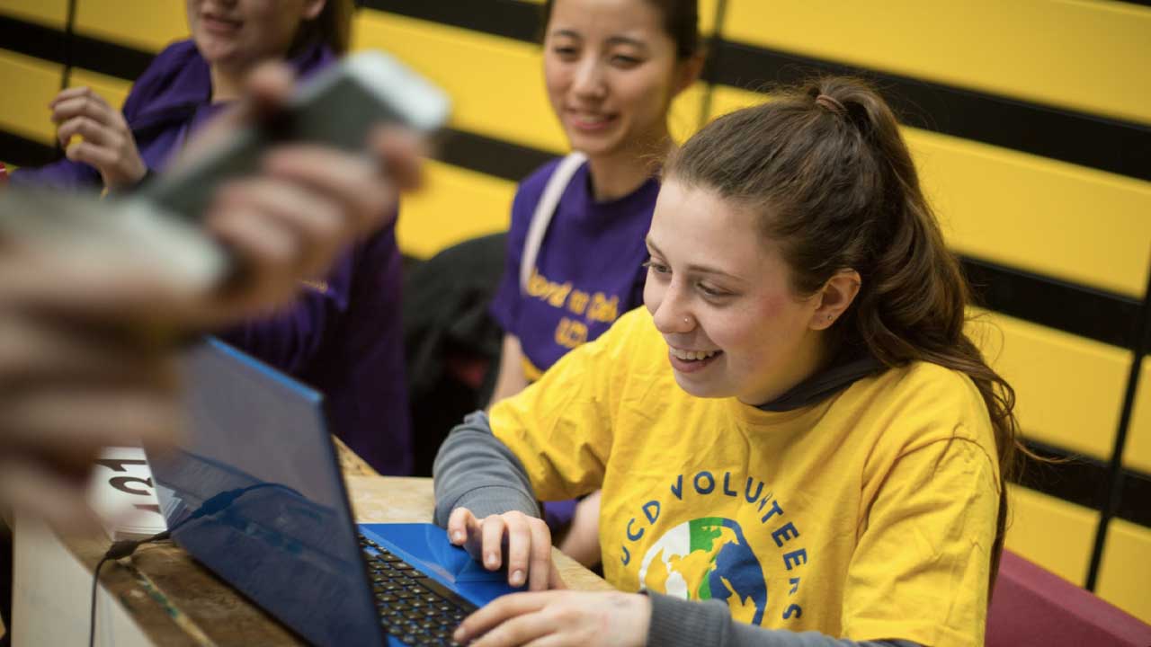 Young woman in yellow T-shirt with UCD VO logo types on a computer while smiling.
