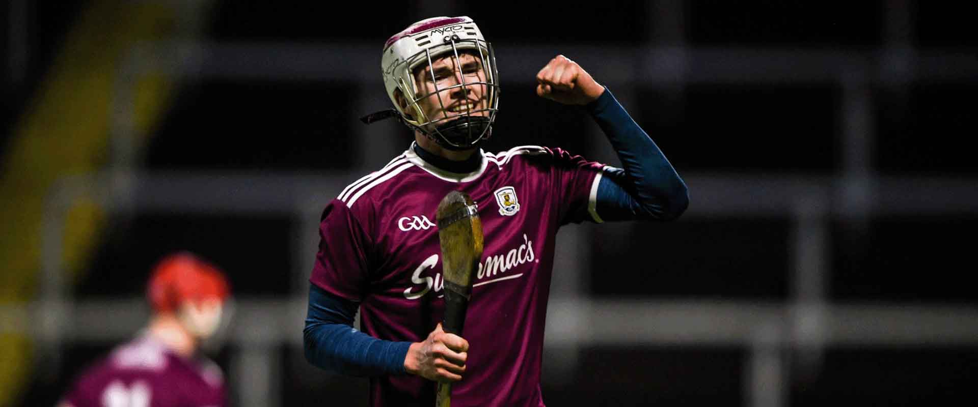 A young man celebrates a goal on a hurling pitch. He is wearing a helmet and a purple jersey.