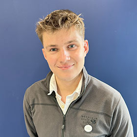 Young main with blond hair and a grey zipped jumper smiles to camera.