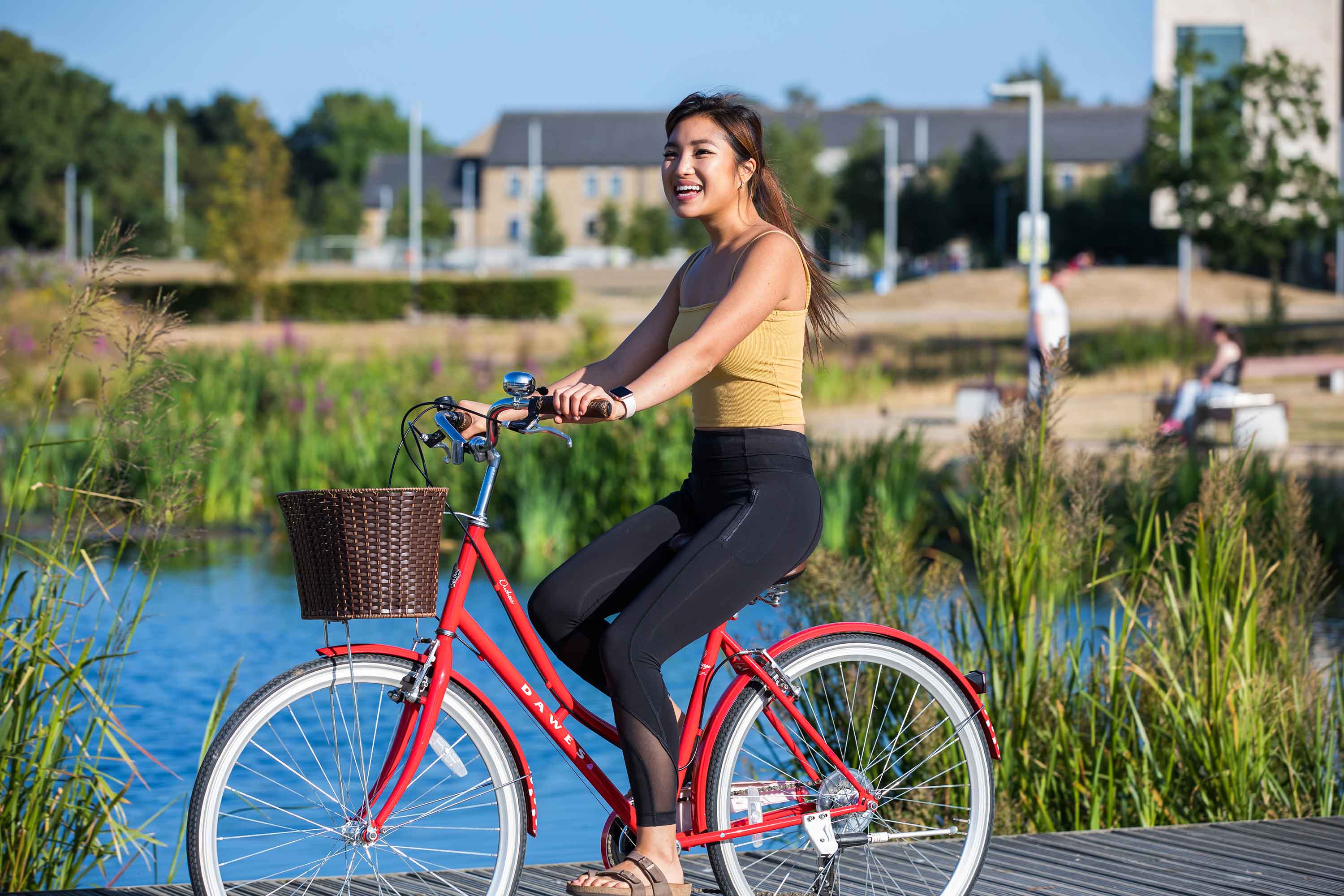 A female student wearing a yellow top cycles a red bicycle by the upper lake at UCD.