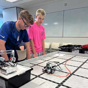 Two UCD students watch a small robot move across a grid-lined mat during a classroom activity.