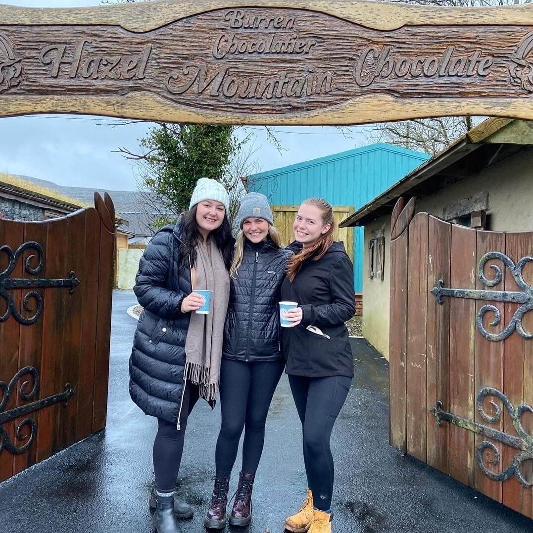 Three young students stand outside a wooden gate. The sign above reads