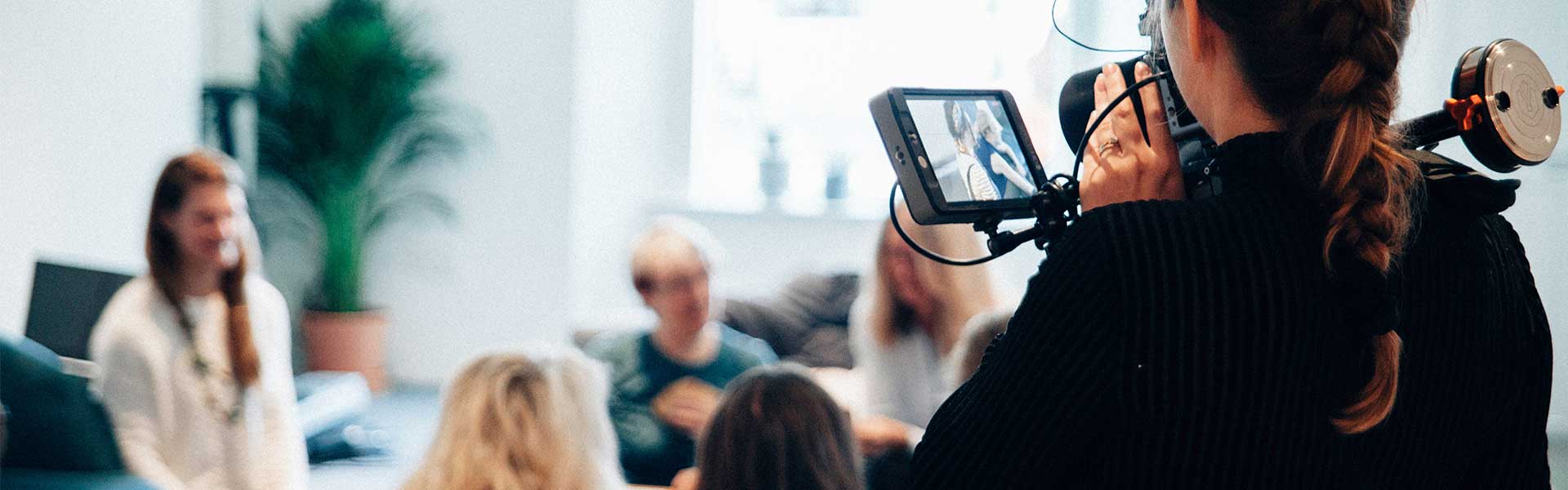 A young woman holds a professional camera, filming a group of people sitting in a white room and chatting.
