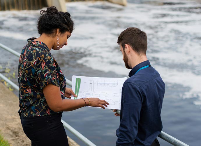 Two people standing by a body of water, reviewing engineering or environmental plans on a large sheet of paper.