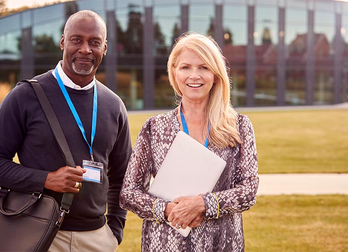 Two people standing outdoors in front of a modern glass building. Both are wearing conference-style lanyards with name badges, suggesting they may be attending an event or working in an academic/professional setting.