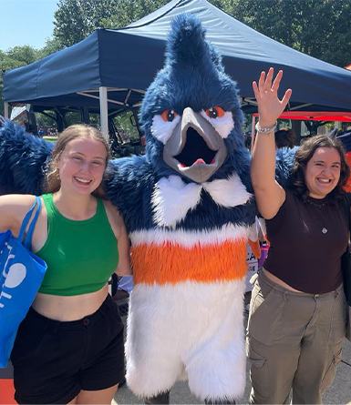 “Two smiling people pose outdoors with a large blue and white bird mascot.