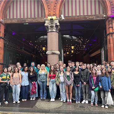 A large group of UCD High School students standing together in front of the entrance to George’s Street Arcade in Dublin, with the red brick archway and hanging lights visible behind them.