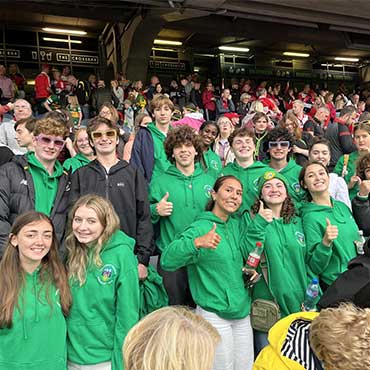 A group of students in matching green UCD hoodies smiling and giving thumbs up while seated in a crowded stadium during a sports event.