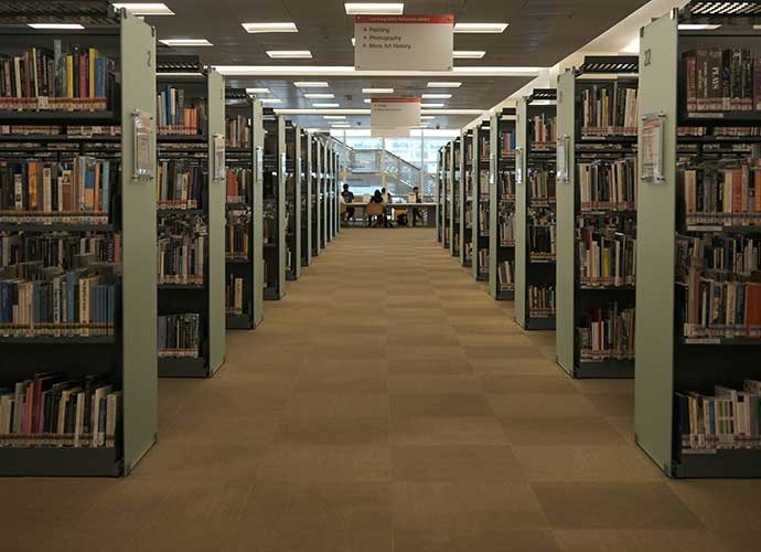 Interior of a library with rows of tall bookshelves on both sides of a wide aisle, leading to a seating area with people reading near large windows.