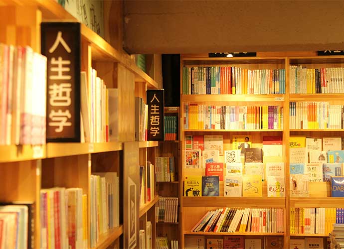 Bookshelves filled with colorful books in a warmly lit bookstore or library, with signs in Chinese characters displayed on the shelves.