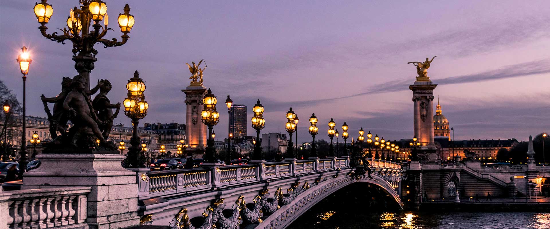 Lanterns line an ornate bridge in the city of Paris with a purple hued sky.