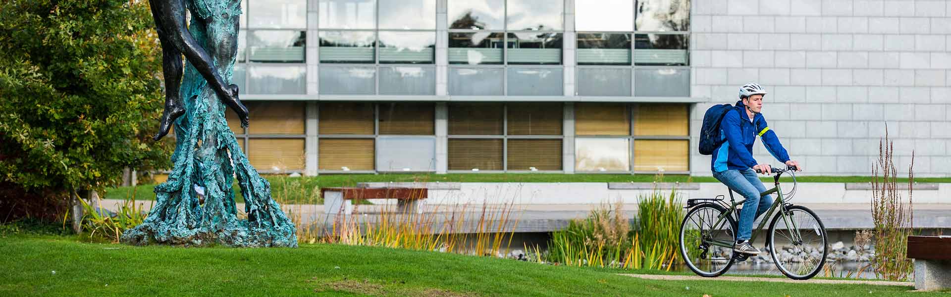 Young man cycles his bicycle past a lake with reads. A grey building with larger windows is in the background.