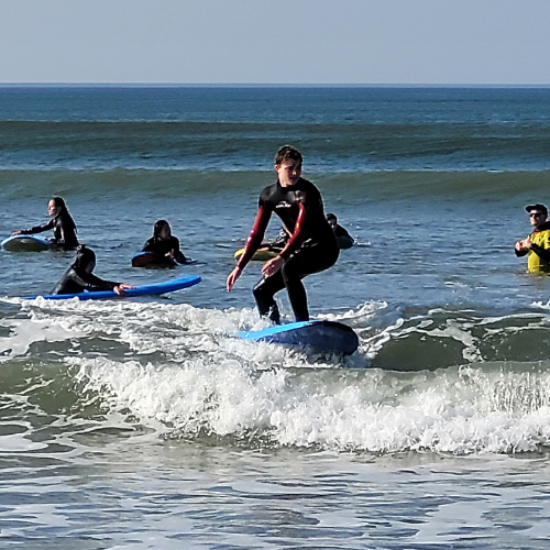 A young man stands a top a surf board surfing a wave. Other surfers are pictured in the distance.