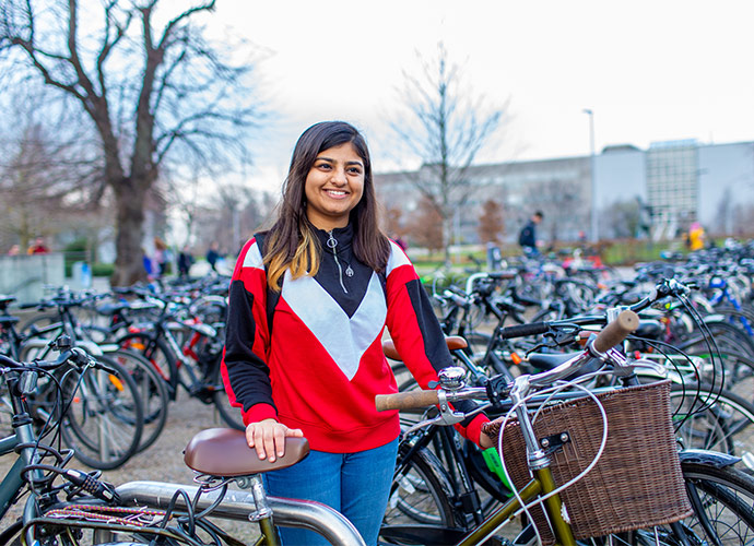 A smiling student stands beside a bicycle with a wicker basket in a busy campus bike parking area, surrounded by rows of bicycles and bare trees on a cloudy day.