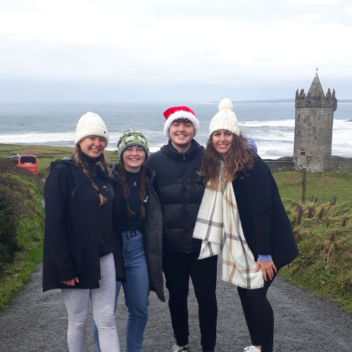 A group of students all wearing hats stand in front of a wild sea and castle.