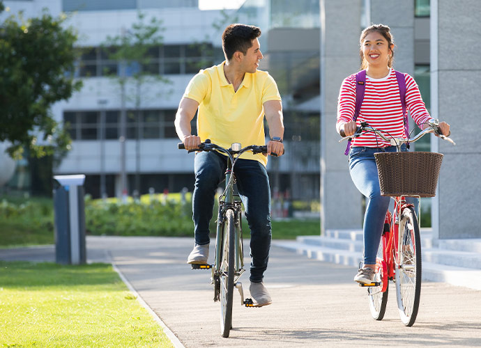 Two students riding bicycles on a university campus, smiling and talking as they cycle along a path on a sunny day.