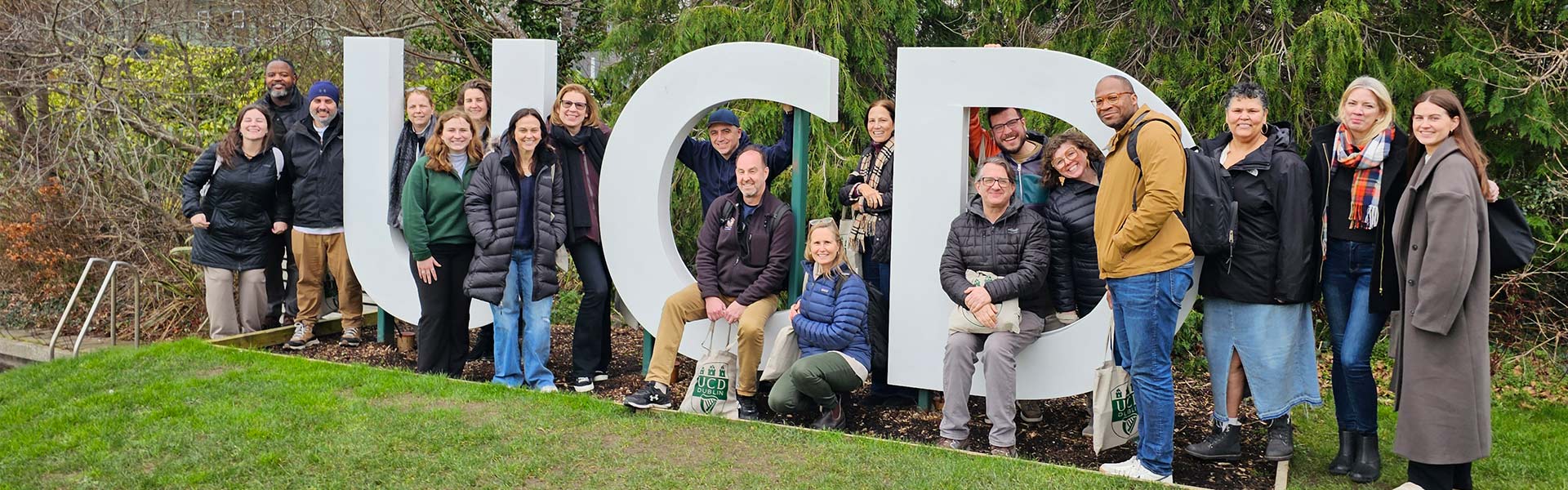A group of people pose for a photo in front of a large UCD sign on the university campus.