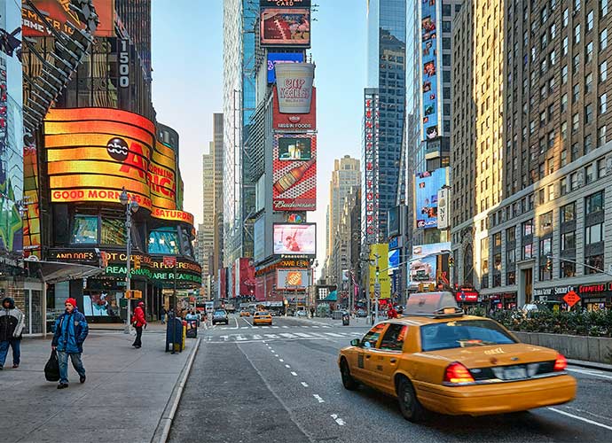 Street-level view of Times Square in New York City, with bright digital billboards, pedestrians on the sidewalk, and a yellow taxi driving through the scene.