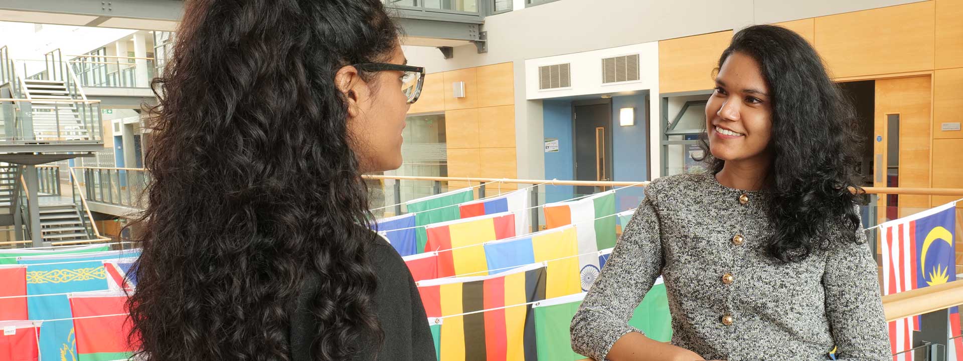 Two female students talking inside a university building decorated with international flags, with modern architecture and staircases in the background.