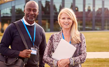 Two university staff members standing outside a campus building, smiling and wearing lanyards.
