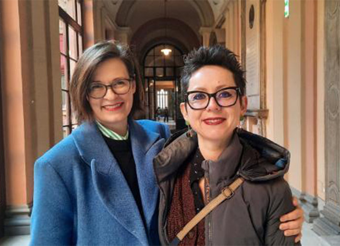 Two woman stand close together smiling to camera.