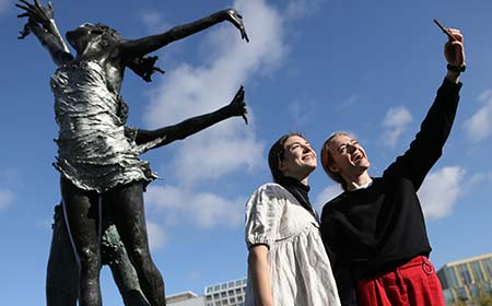 Two students take a selfie with large bronze statues dancing.