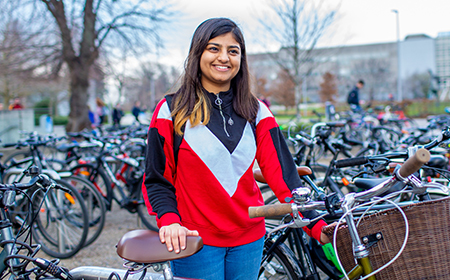 A young woman stands holding a bicycle in front of bicycle racks on a grassy campus.