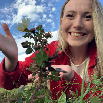 A woman smiling outside wearing red blazer on the ground with a flower with white petals in the foreground. The blue sky with several clouds can be seen in the background.