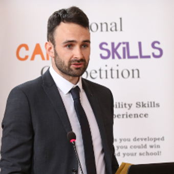 A half body shot of a man in the suit looking off to the side with a white poster in the background relating to language learning