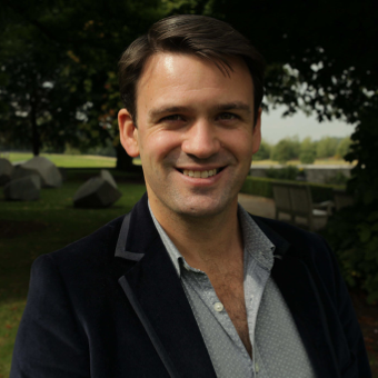 A portrait of a man smiling looking into the camera wearing a gray shirt and dark blazer with grass and shaded tree in the background