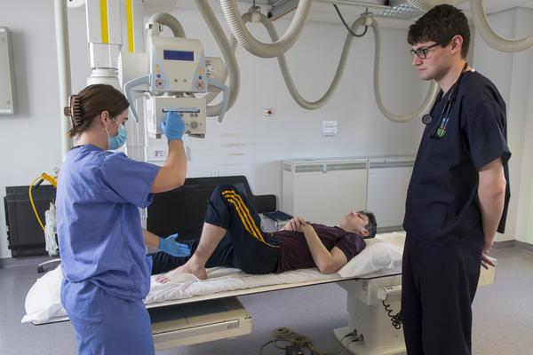 Person lying on bed attached to radiography equipment, student looking at person in scrubs using the equipment