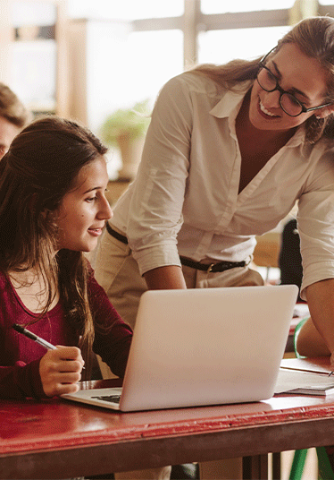 Female teacher with long blonde hair and glasses leaning on student desk helping with her work.