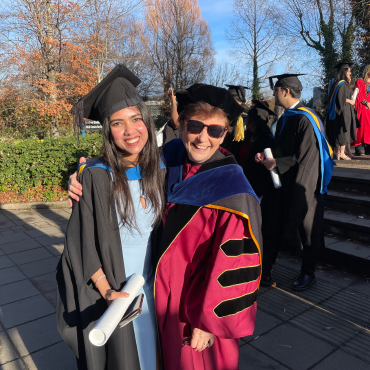 A graduate and an academic in caps and gowns outside on a sunny day.