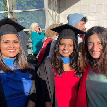 UCD graduates and UCD academic in caps and gowns outside on a sunny graduation day.