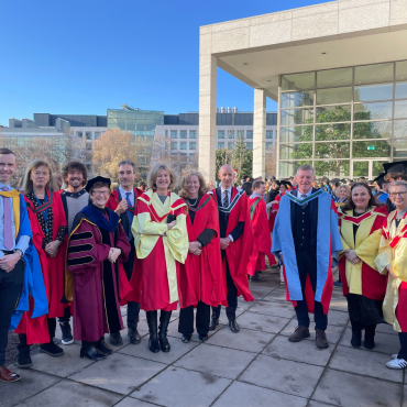 UCD academics in caps and gowns outside on a sunny graduation day.