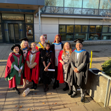 UCD graduates and UCD academic in caps and gowns outside on a sunny graduation day.