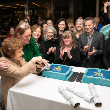 Woman cutting a cake surrounded by a group of people at the UCD Physiotherapy 70th anniversary celebration