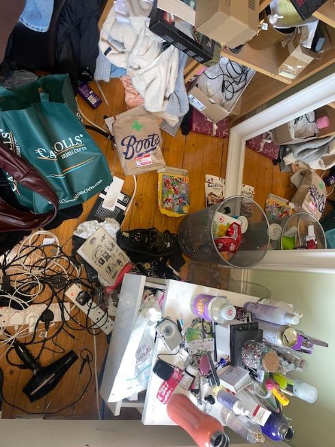 A cluttered room with clothes, cables, shopping bags, and various items scattered on the floor. A small table is piled with beauty products, a hairdryer, and makeup. A mirror leans against the wall amidst the mess.