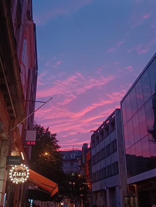 A vibrant city street at sunset with pink and purple clouds. Buildings line the street, including a restaurant with a Zizzi sign. A digital clock on a building displays 18:59. Trees and a bird silhouetted against the colorful sky.