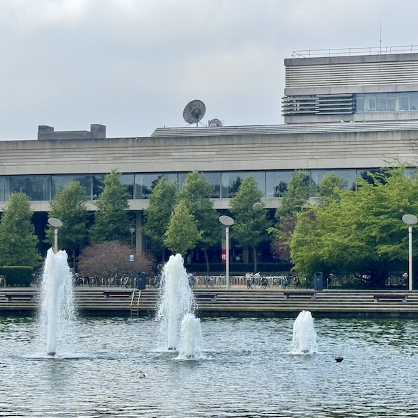 3 fountains in a lake with some trees and a brutalist concrete building behind it