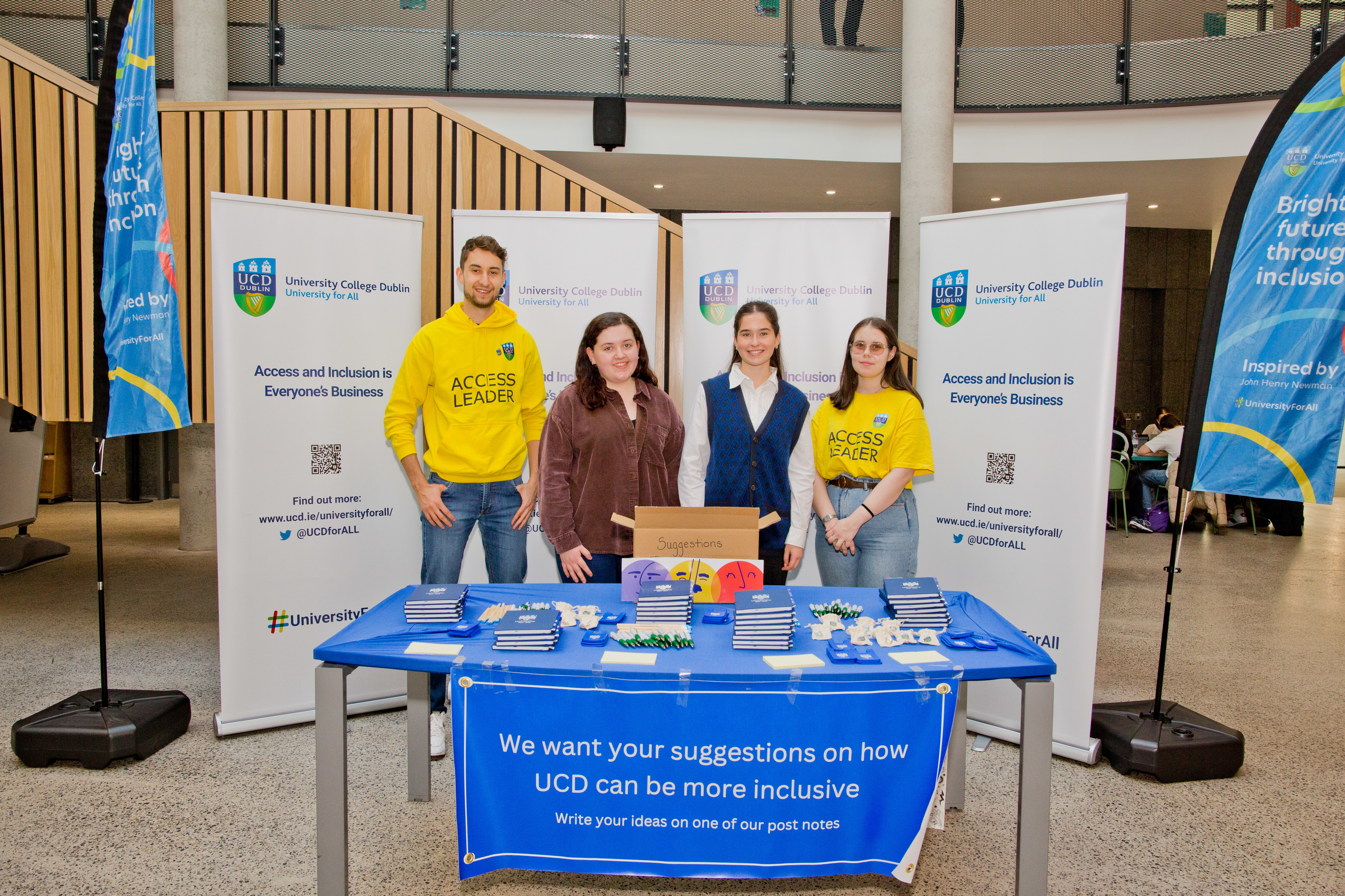 4 students standing in front of a blue table. The table has a sign at the front, the table has merchandise and donuts on it. It has 4 white popups at the back.