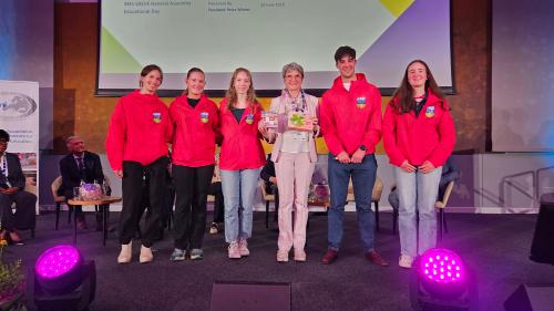 Group photo of student volunteers at the 38th EAEVE General Assembly 2025. From left to right: Laura Zopf, Amy Beechinor, Isla Wilson, Prof. Dr. Petra Winter (President of EAEVE, holding a thank-you gift), Eoin O'Toole, and Hannah Lynam. All students are wearing red UCD jackets and smiling on stage. Not pictured: Student volunteer Rebecca Stevenson. A presentation screen and seated attendees are v