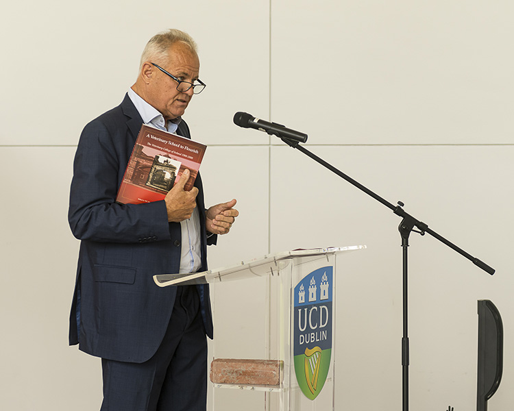 Professor Michael Doherty speaking to guests from the podium in the O'Reilly Hall Conservatory, holding a book in his hand